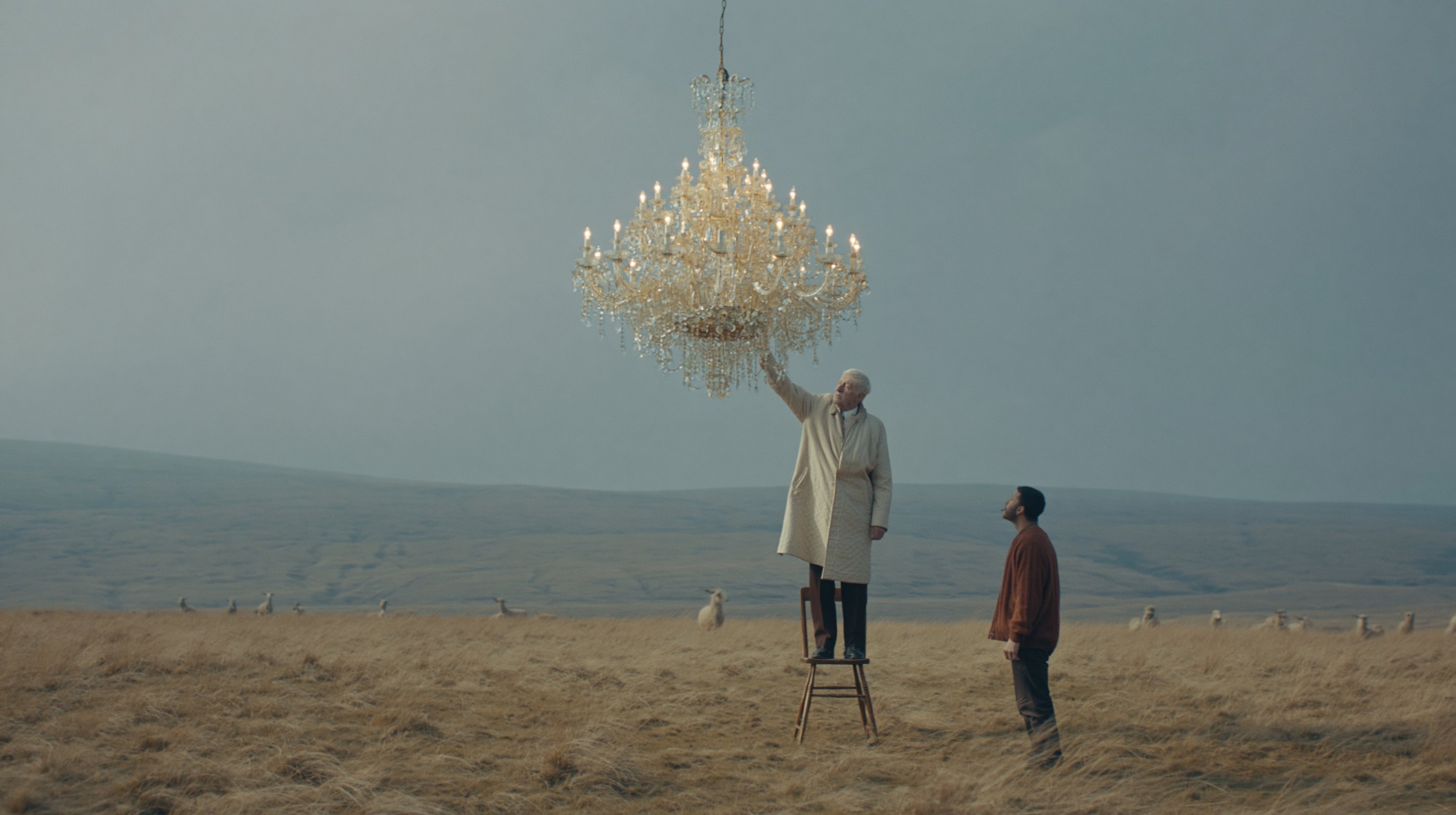 An older man in a pale coat stands on a chair and reaches towards a chandelier while a younger man watches in a field.