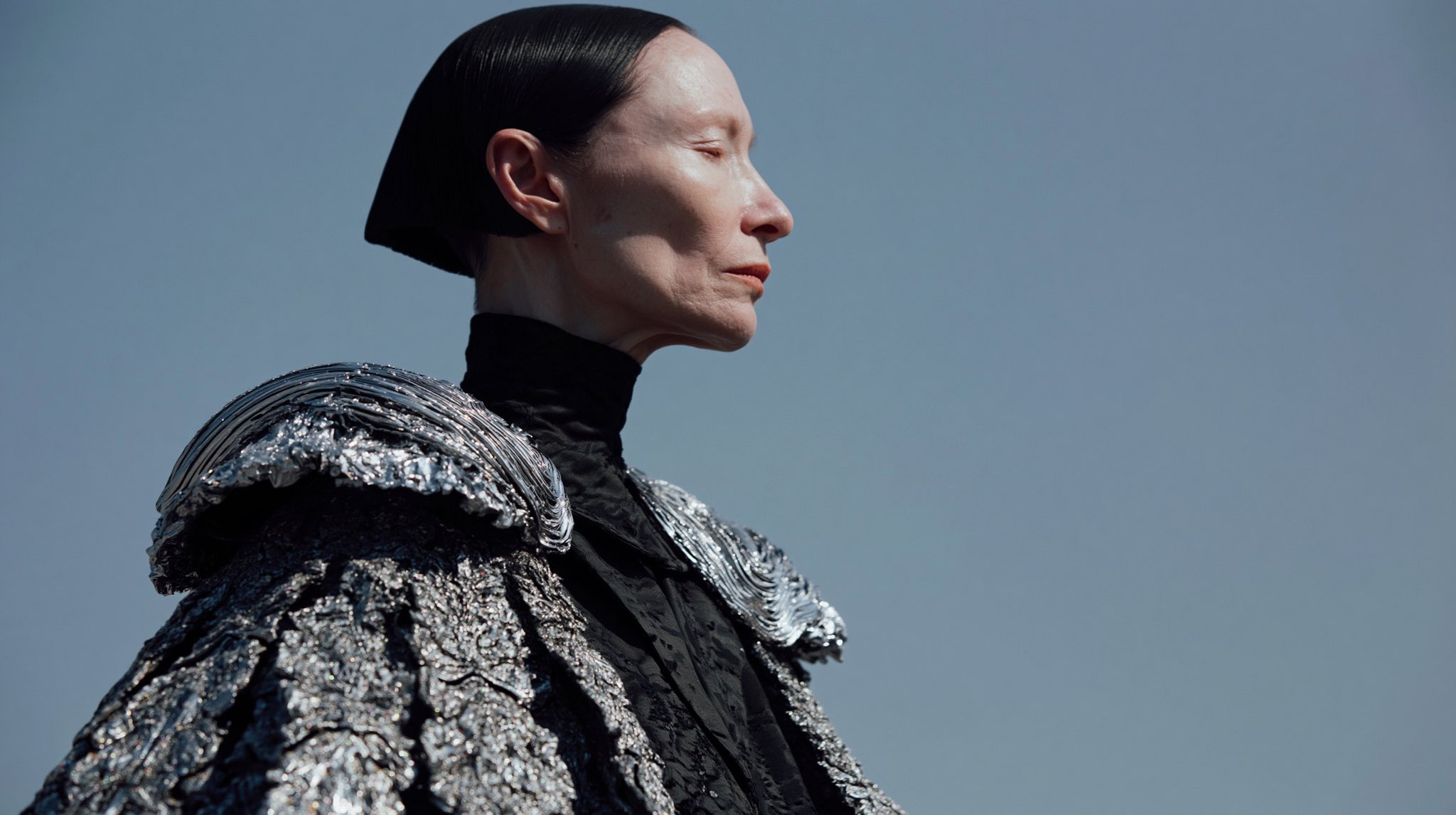 Profile portrait of a pale female Council member wearing a black high-necked garment and textured silver shoulder piece.