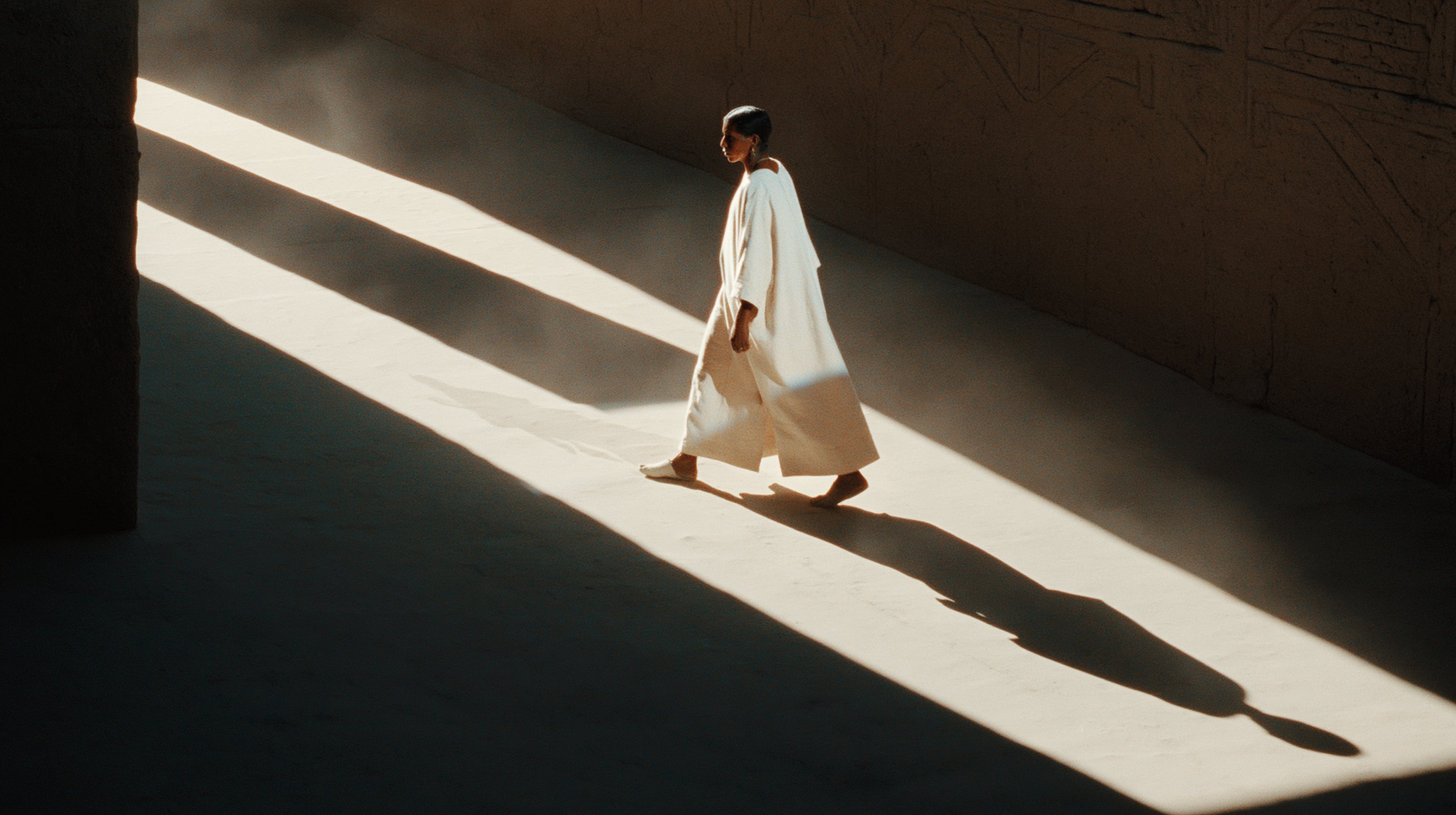A pale-robed figure walks through a narrow stone passage crossed by hard bands of sunlight and shadow.