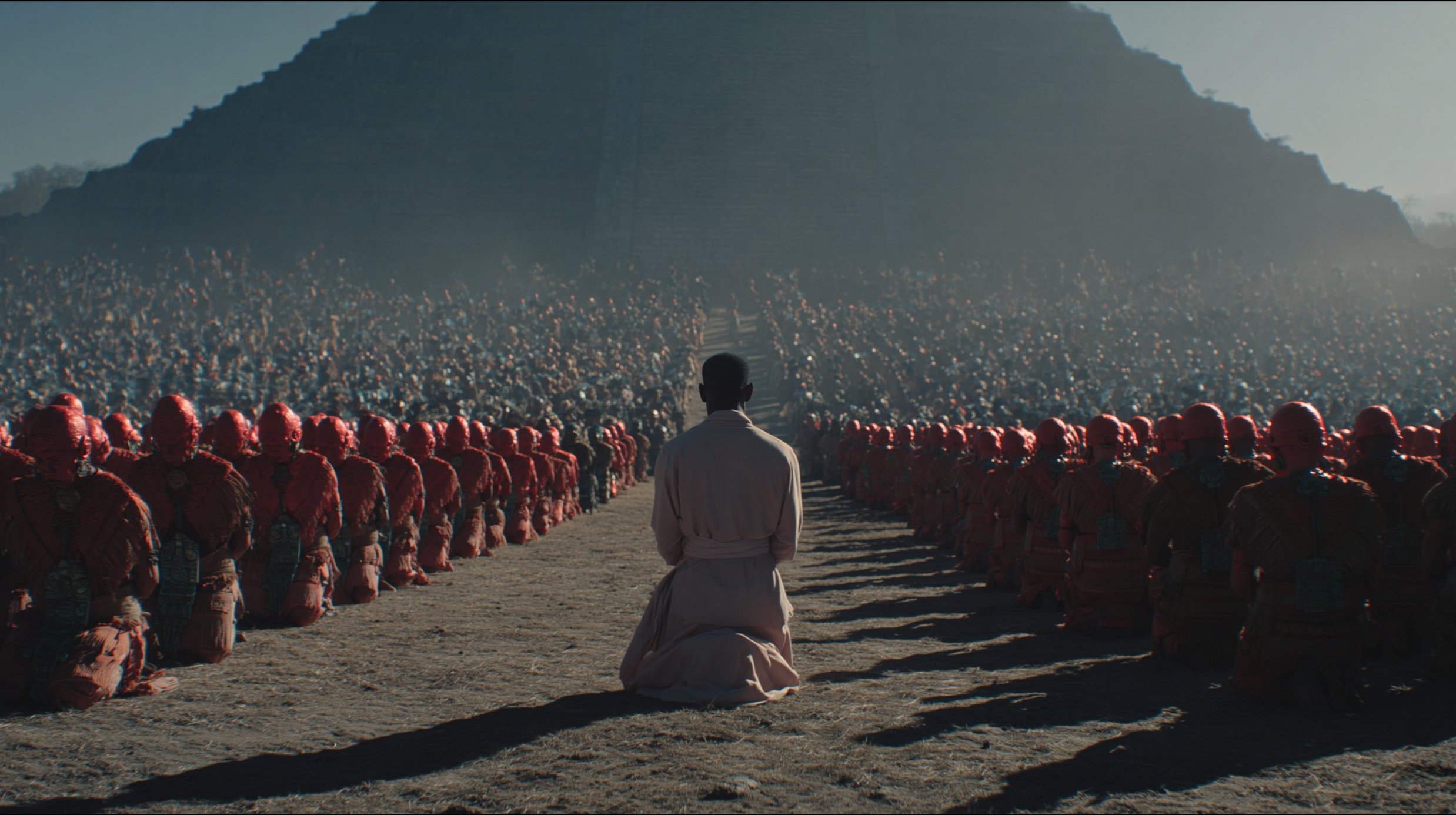 Rows of red-robed figures kneel in synchronised formation before the pyramid, viewed from behind a solitary standing figure.