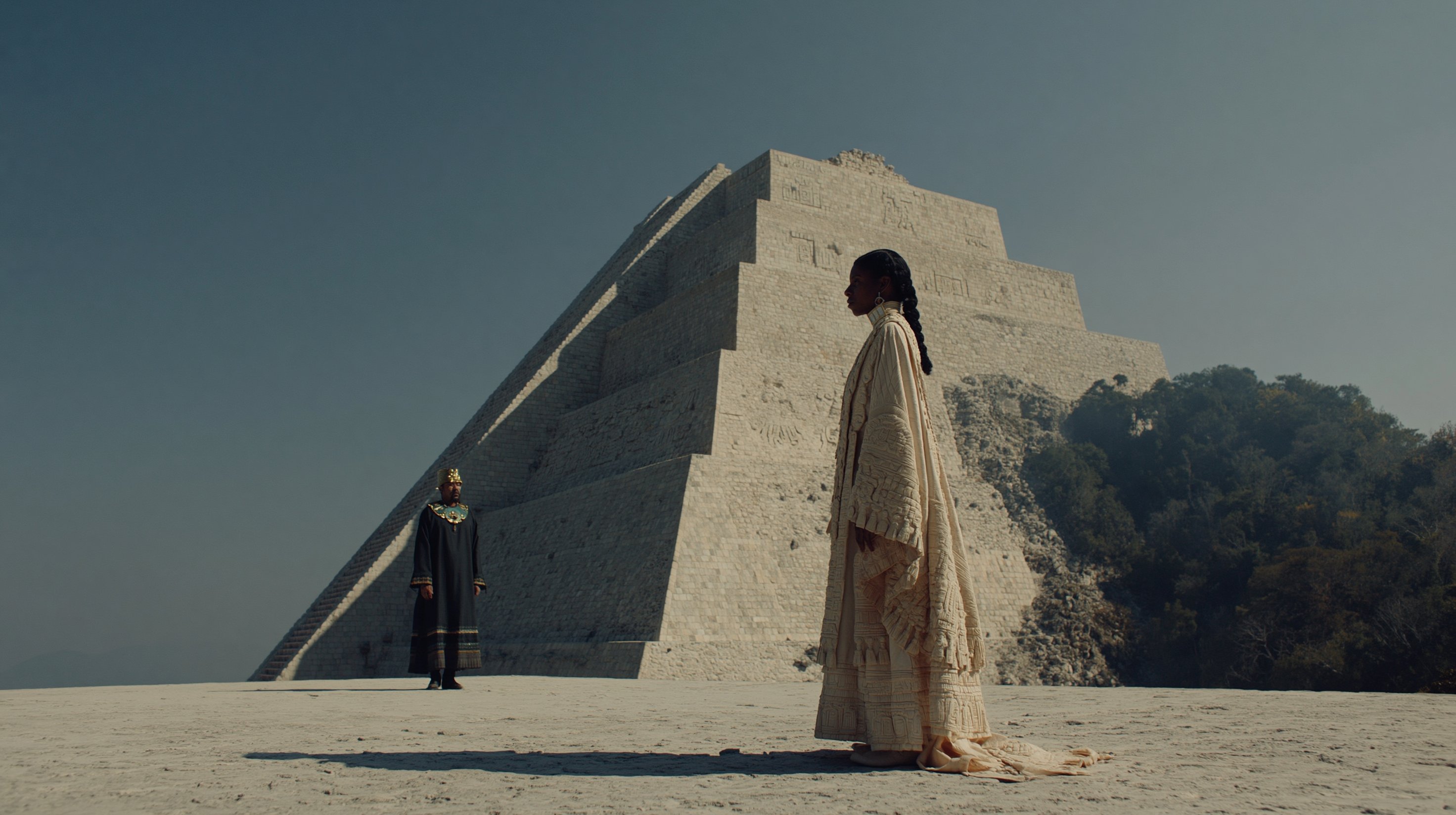 An emissary in pale ceremonial robes faces a dark-robed leader across a sunlit pyramid platform.