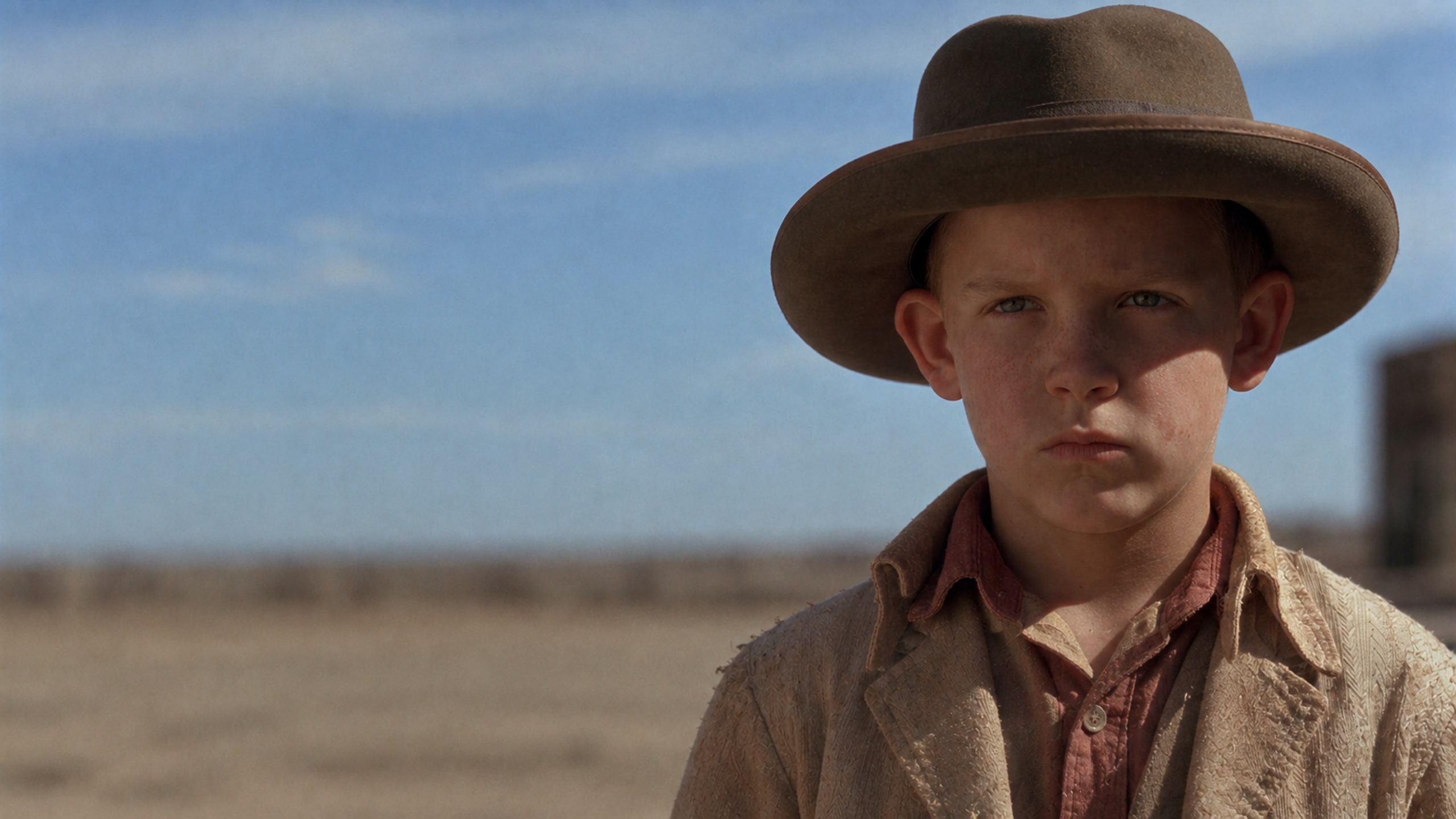 Close-up of a small boy in a worn tan hat and dusty jacket, standing alone in the desert.