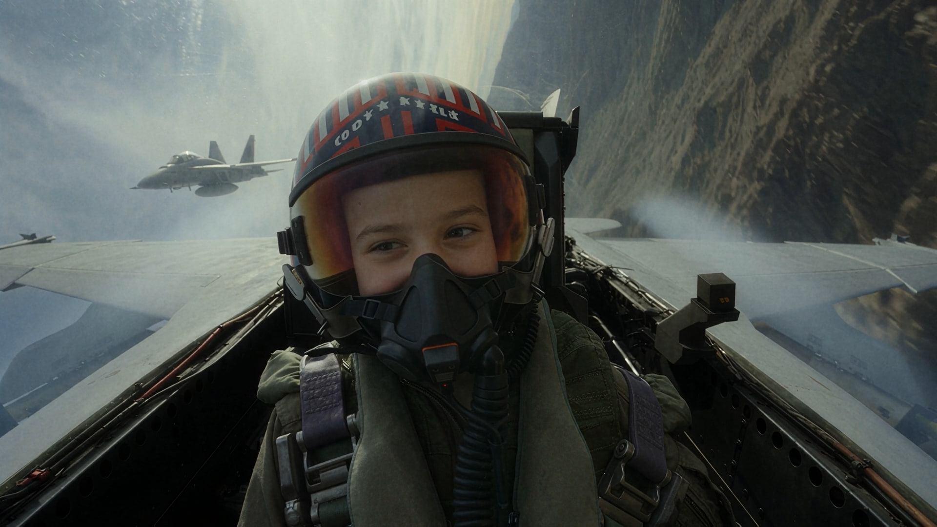 A child fighter pilot in a helmet and oxygen mask looks back from the cockpit as a second jet follows through the canyon.