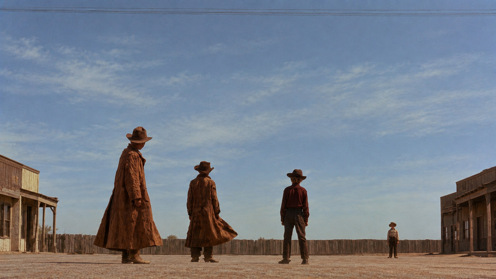 Three children in long coats and cowboy hats face one small child across a dusty Western street under a wide blue sky.